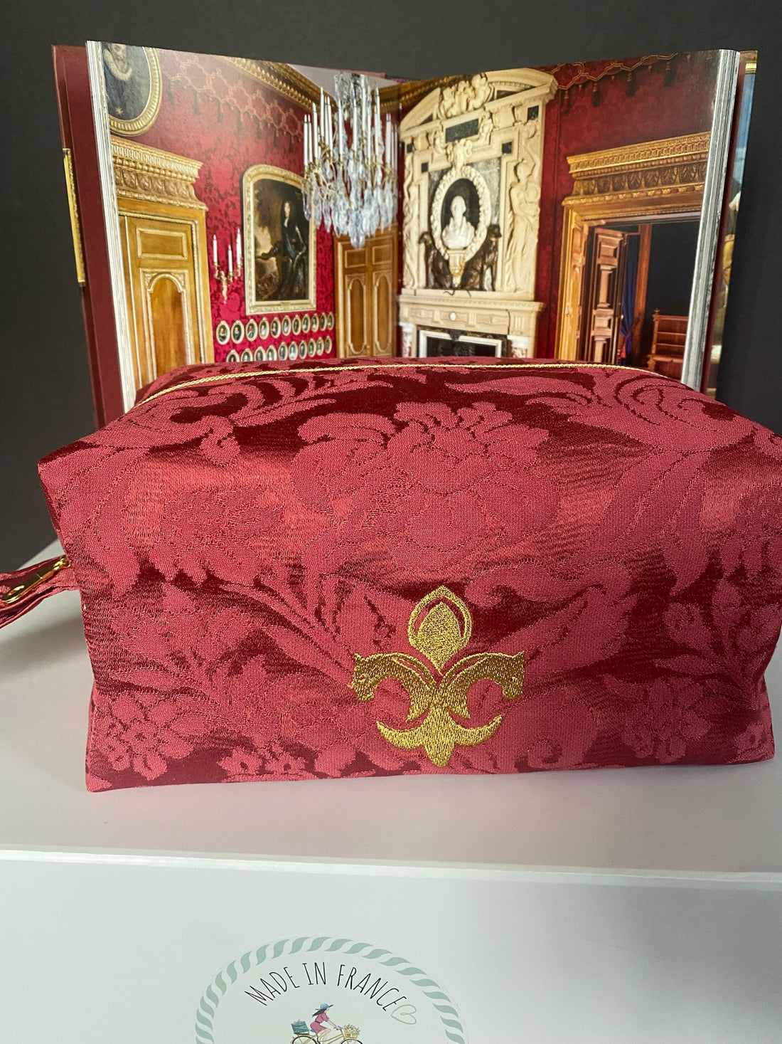 Red damask washbag embroidered with a gold fleur-de-lis, against the décor of a château salon